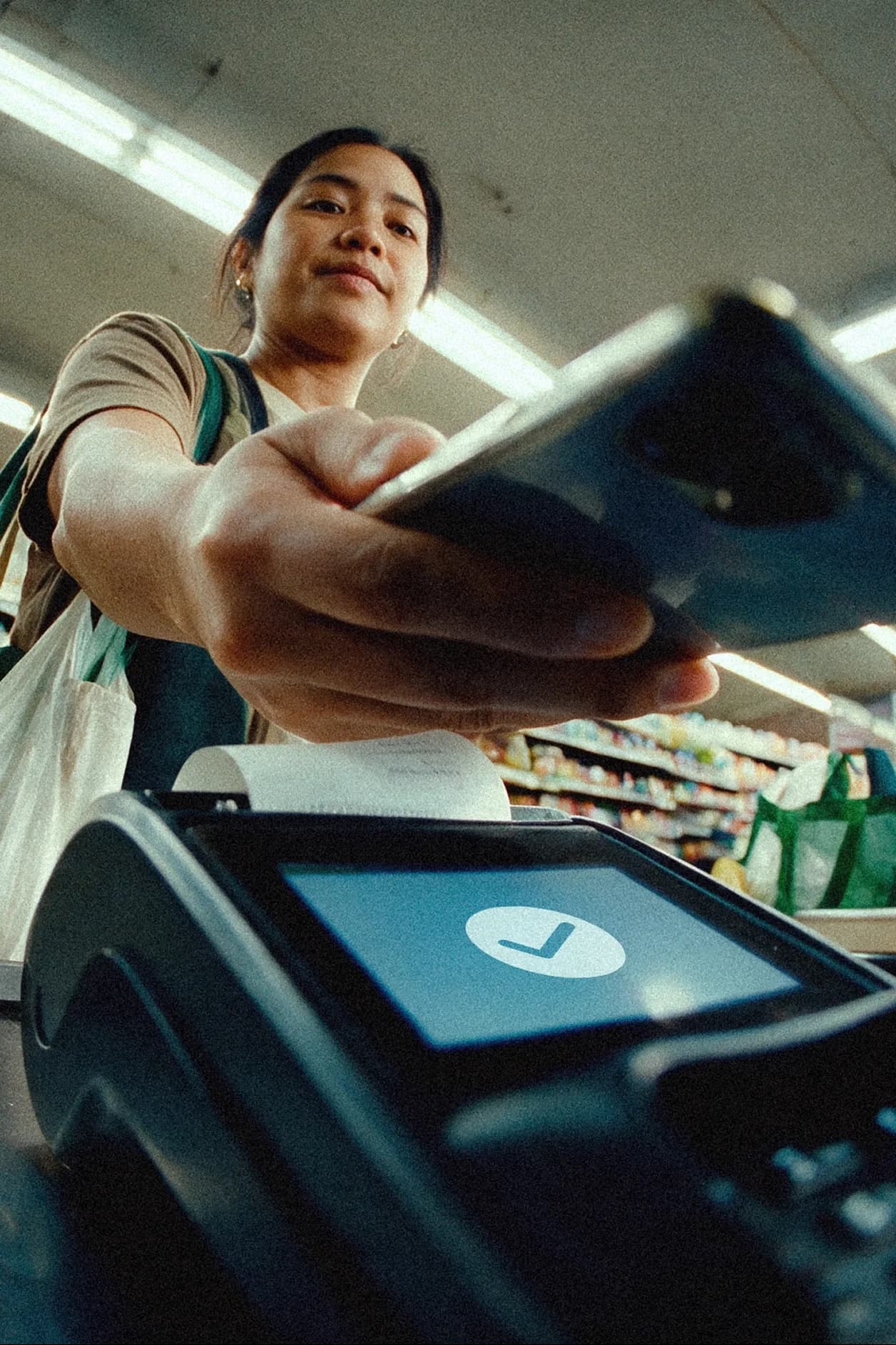 Woman at a grocery checkout tapping her phone on a payment terminal that shows a successful checkmark, with reusable bags on her shoulder.