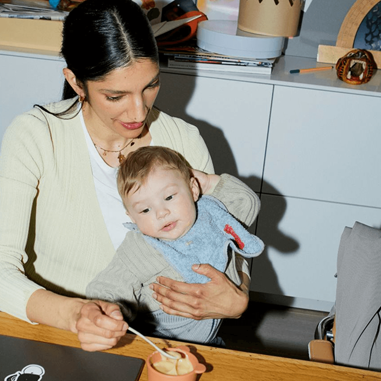 Woman holding a toddler at a kitchen table while feeding the child from a small bowl.