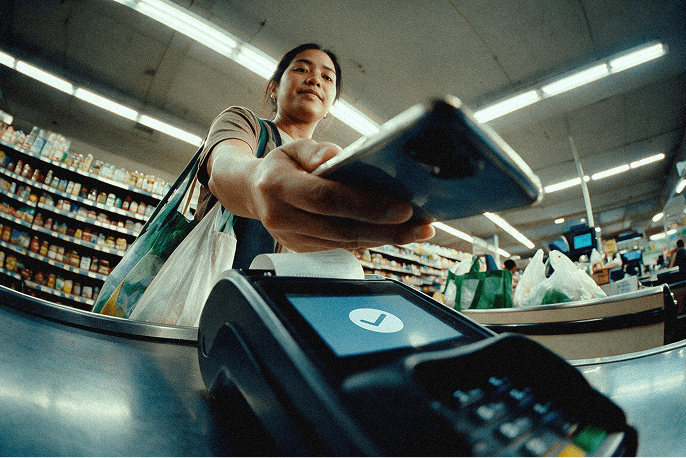 Woman at a grocery checkout tapping her phone on a payment terminal that shows a successful checkmark, with reusable bags on her shoulder.