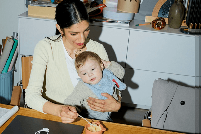 Woman holding a toddler at a kitchen table while feeding the child from a small bowl.