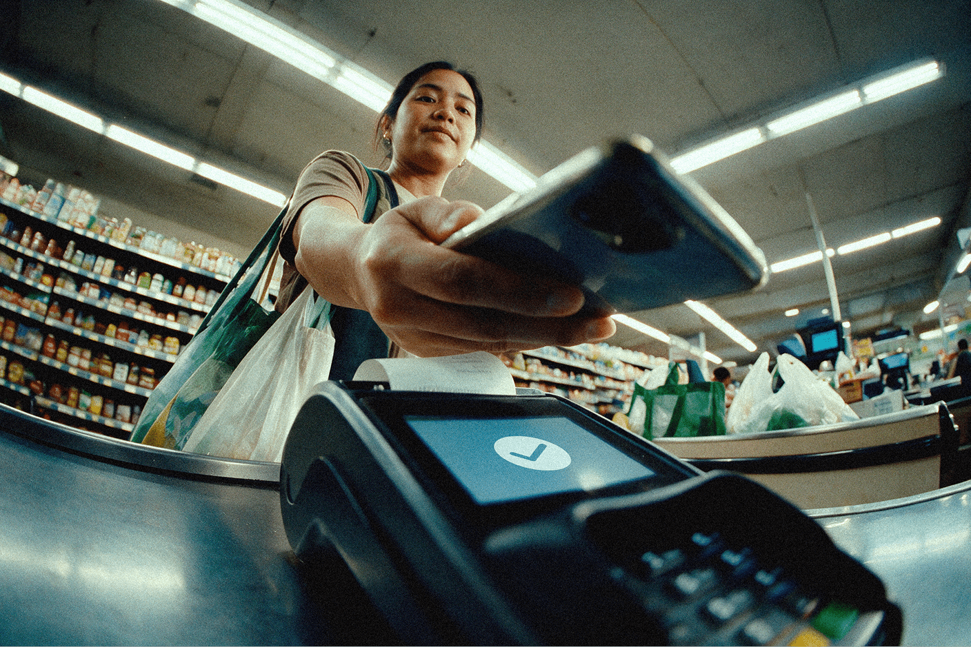 Woman at a grocery checkout tapping her phone on a payment terminal that shows a successful checkmark, with reusable bags on her shoulder.