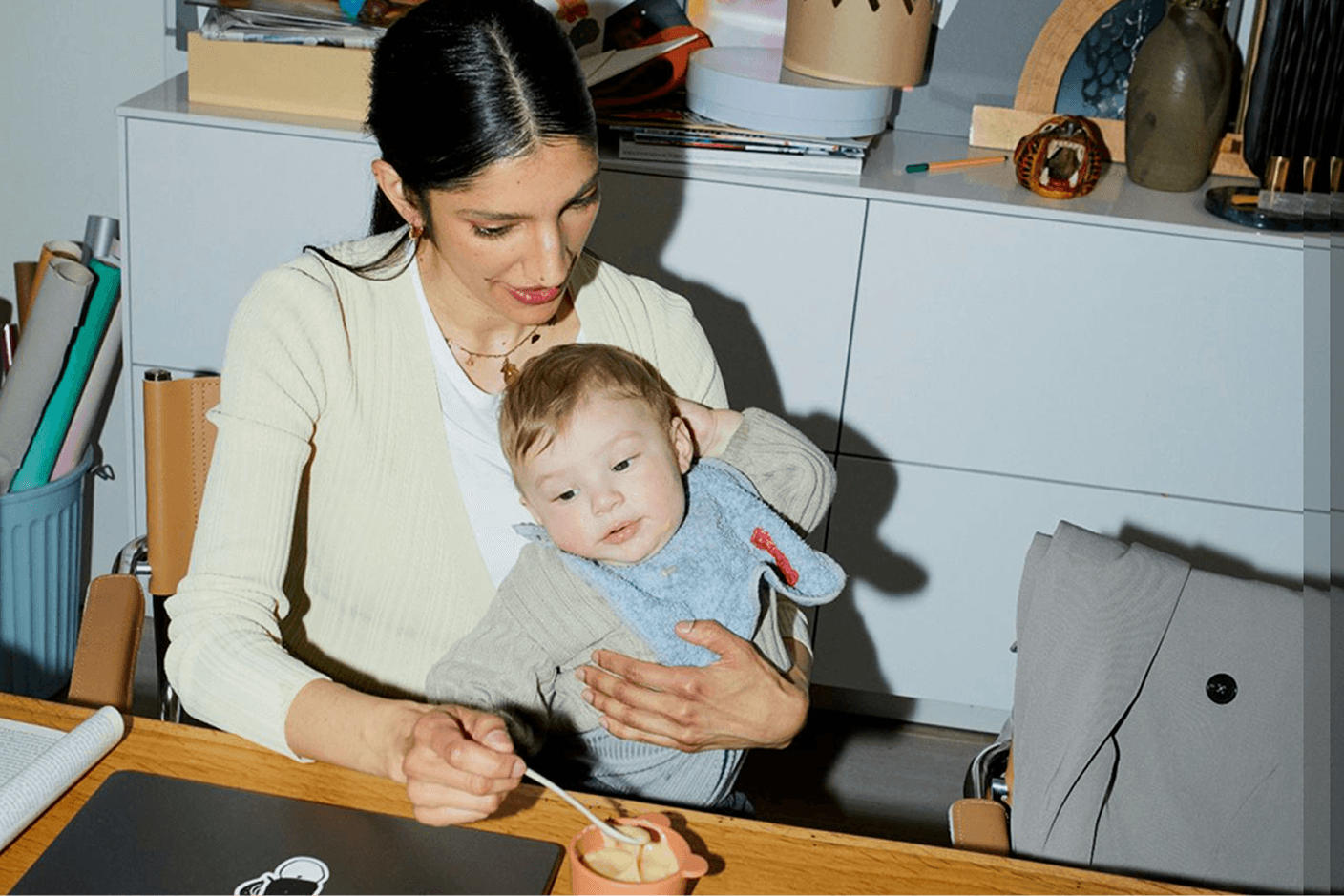 Woman holding a toddler at a kitchen table while feeding the child from a small bowl.