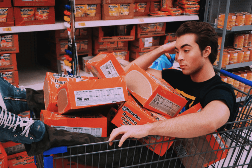 A Canadian man who went to the store to buy just one thing, sits in a grocery cart filled with on-sale soup boxes. He's wondering about his impulse buys.