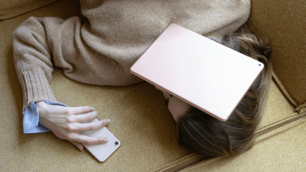 An unemployed person laying on the couch with their phone and tablet, contemplating whether they can get a credit card without a job