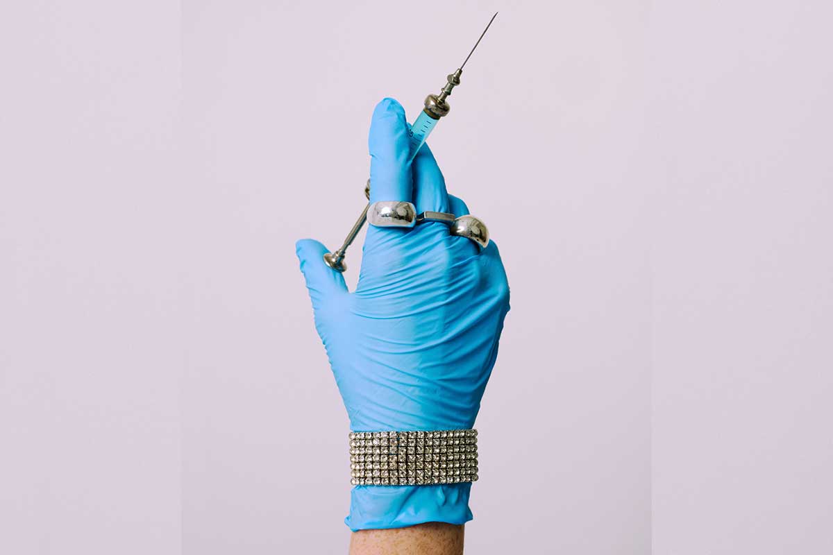 A Canadian hand with a needle, dressed in jewellery, to suggest the vanity of biohacking better bodies with peptides.