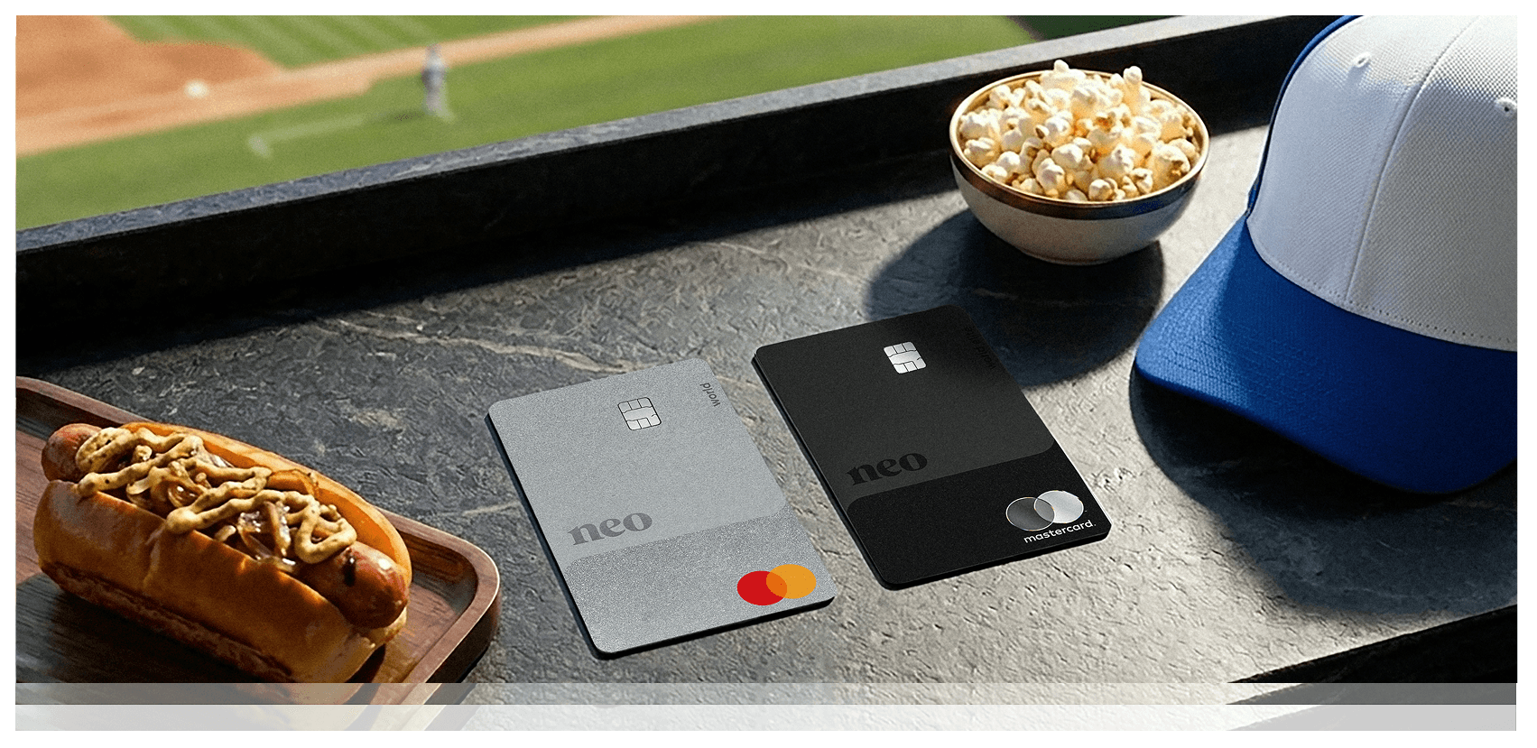 Two Neo cashback cards on a ledge at a baseball stadium. On the left is the Neo World Mastercard - Everywhere, on the right is the matte black Neo World Elite Mastercard - Everywhere next to a bowl of popcorn and a blue and white baseball cap.