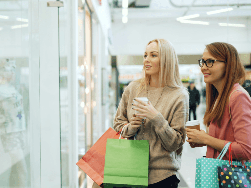 Two Canadian women looking at a store window, with shopping bags in arms, wondering if it's a better deal to shop on Black Friday or wait until Boxing Day.