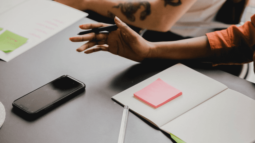 Two people mid-conversation by a desk with a phone, laptop and sticky notes, chatting about how to calculate interest on your savings account in Canada.