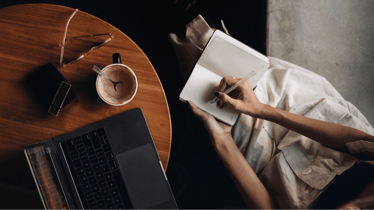 A person sitting by a wooden table, drinking a cup of coffee and writing in a notebook, doing research on debt consolidation in Canada