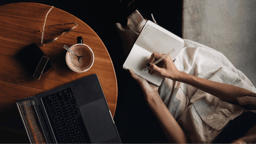 A person sitting by a wooden table, drinking a cup of coffee and writing in a notebook, doing research on debt consolidation in Canada