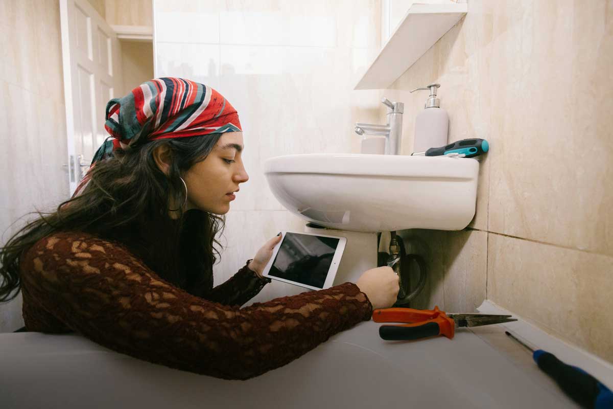 A Canadian woman fixing a leak in her bathroom, with the help of a how-to video, while also trying to decide if she needs to hire someone for home repairs.
