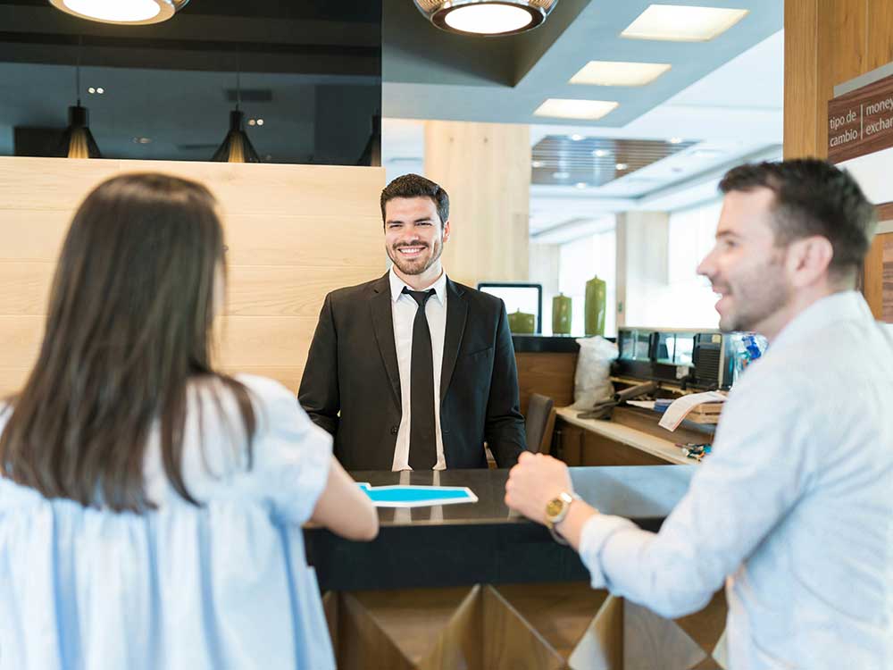 Two Canadians checking into a hotel, unaware of the hidden fees they might be paying (among other fees, from banking to investing).