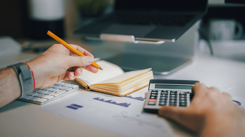 A person's view of their desk, where they have a notebook, calculator and paperwork with graphs and charts, as they attempt to figure out how interest works on a savings account in Canada.