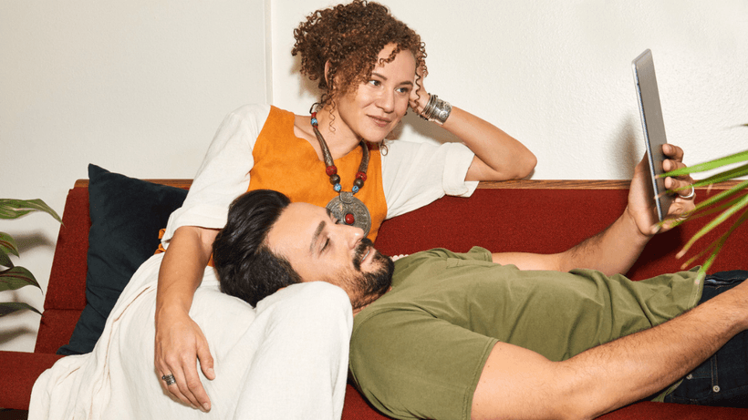Two people lounging on a sofa. One person rests their head on the other's lap while holding up a digital tablet with information about joint account tax implications in Canada
