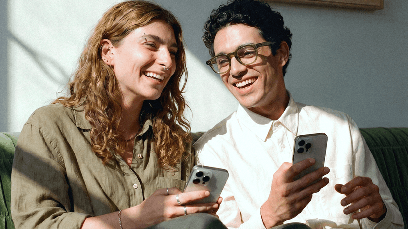 A happy couple sitting on a sofa and looking at their smartphones, illustrating how to manage a joint bank account in Canada.