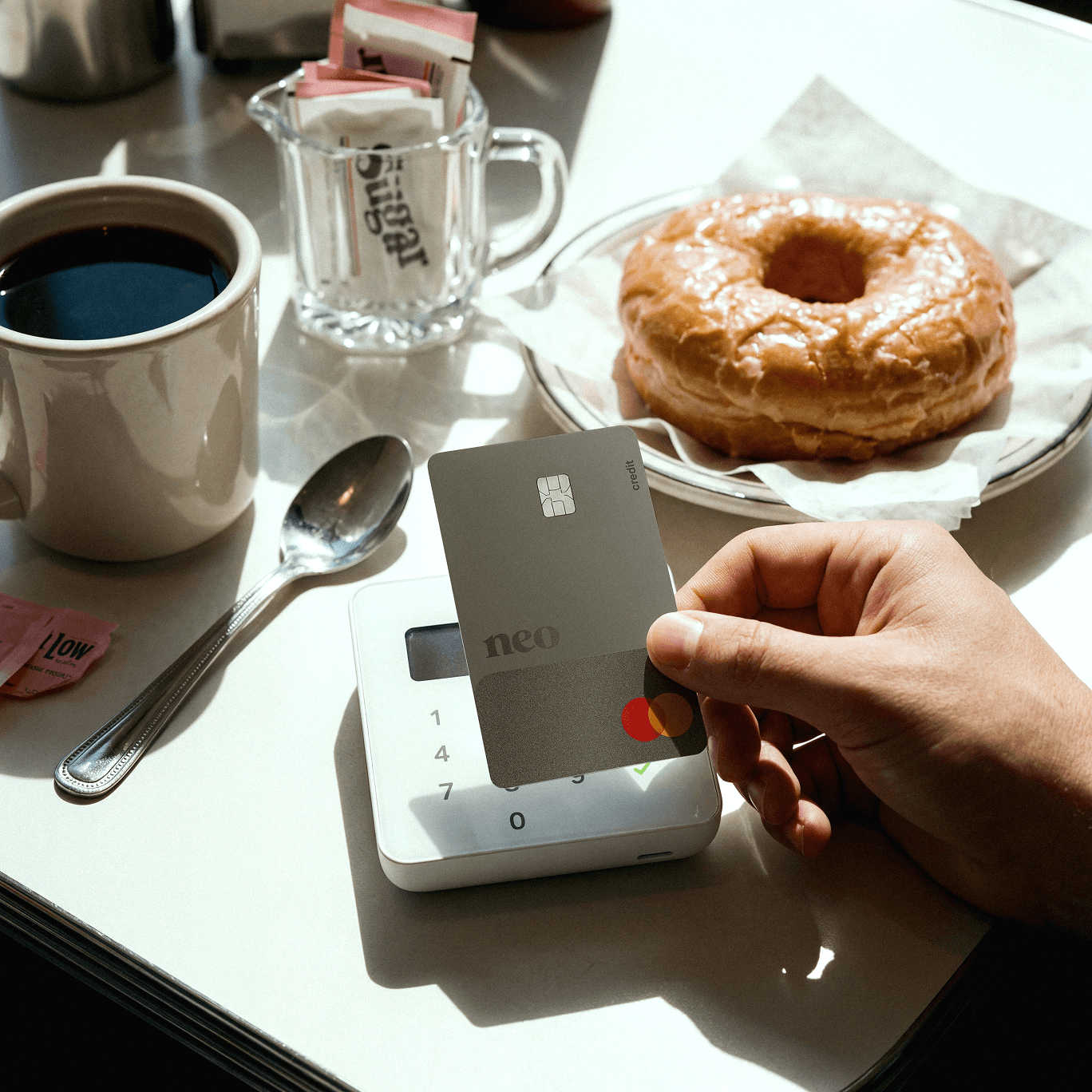 A person uses a Neo Mastercard, an example of a top secured credit card in Canada, to pay for a meal. They hold their Neo secured credit card over a payment terminal on a surface, which also has a cup of coffee and a donut.