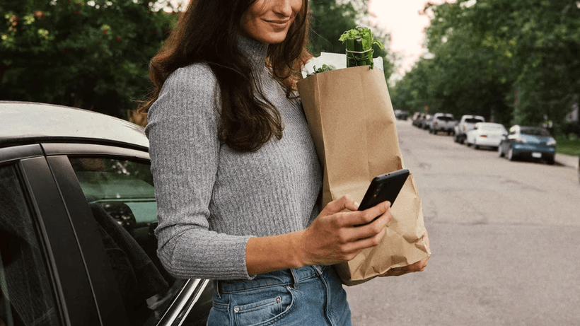 A woman leaning against a black car on a residential street, holding a brown paper grocery bag in one arm while checking her smartphone.