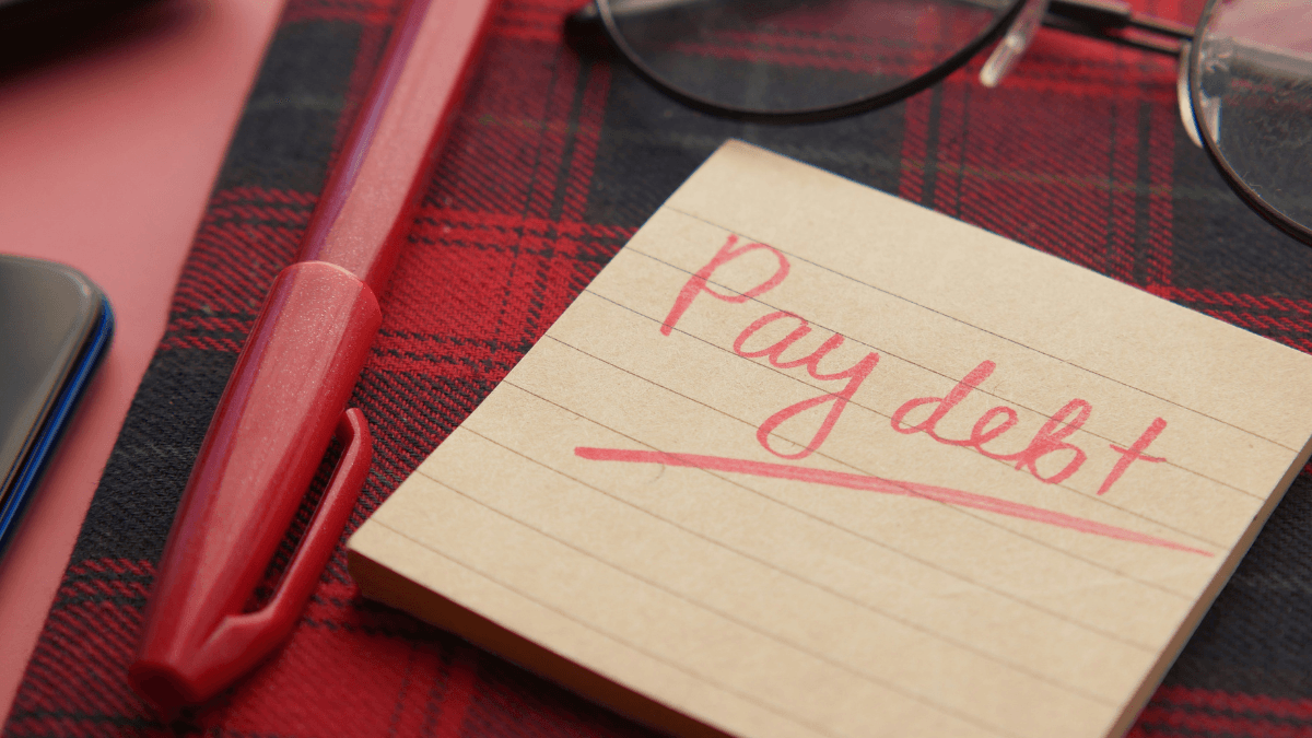 A notepad on a red tartan surface beside a pen and a pair of glasses. The notepad reads, "Pay debt".