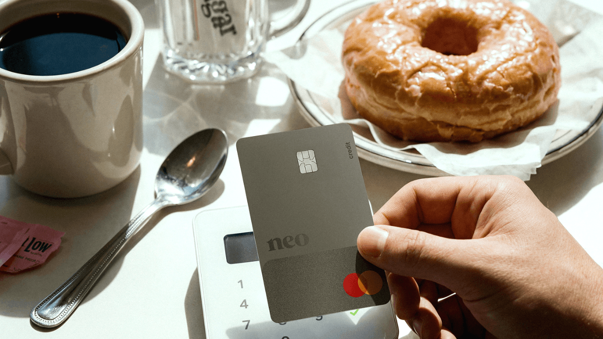 A person's hand holding a gray Neo credit card over a white payment terminal at a restaurant, illustrating the everyday purchasing power of a credit limit increase.
